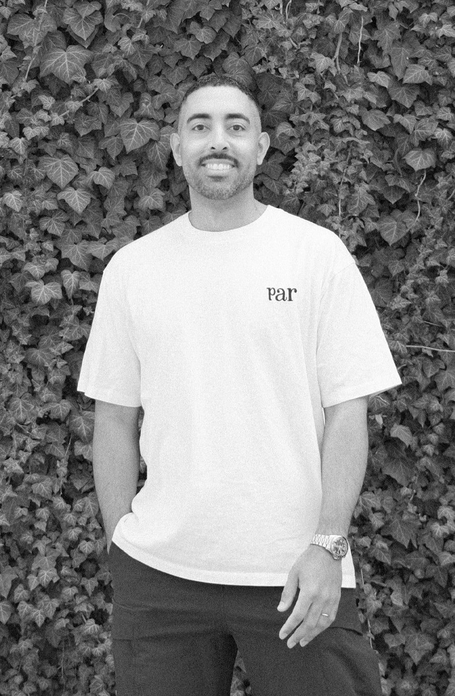 Black and white headshot. White shirt, in front of a green vine wall.
